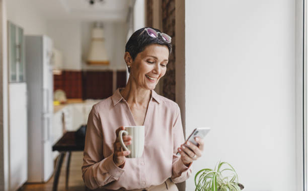 istock Gorgeous elegant mature caucasian brunette lady drinking coffee with mug in hands, browsing social media on smartphone or chatting online standing next to window with kitchen on background 2174090598