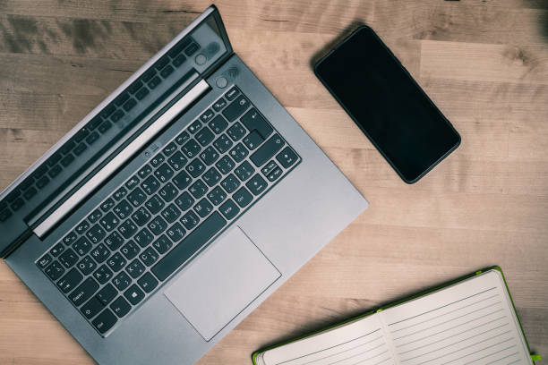 An overhead shot featuring a laptop, a cellphone, and an open notebook arranged on a rustic wood table. The laptop is silver and slightly angled, showcasing its keyboard and screen. The cellphone is black, lying next to the laptop, displaying a home screen. The notebook, with a green cover, is positioned beside the other devices, adding a personal touch. Soft natural lighting highlights the textures of the wood table, creating a warm and inviting workspace atmosphere.