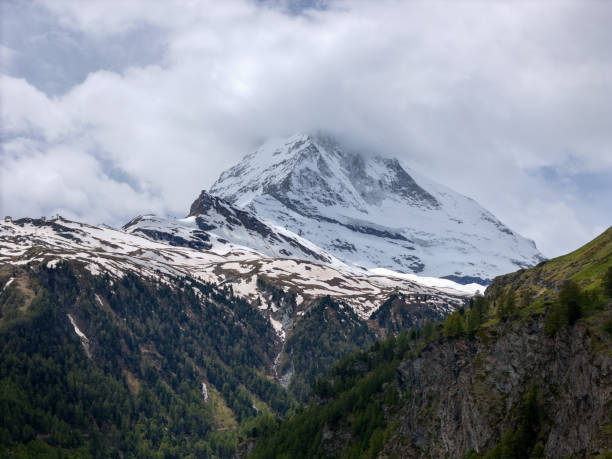 A towering snow-capped mountain partially shrouded in clouds, surrounded by lush green forested slopes, showcasing the beauty of nature.