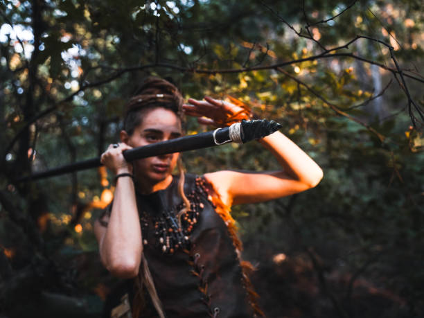 A warrior woman posing powerfully with her staff in a dense forest, her costume adorned with feathers and tribal elements.