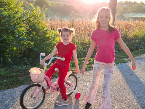 Two happy girls having fun while running and riding a bicycle through village road during late summer day