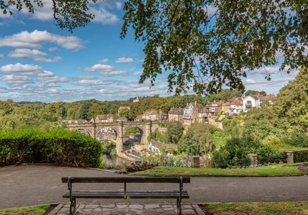 A 19th Century built viaduct crosses a deep valley surrounded by woodland. Buildings line the hillside and a church tower is on the skyline. A bench is in the foreground.