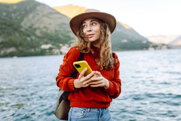 istock A young woman using her phone while sitting by the waterfront in a scenic mountain town during a sunny day. 2174122347
