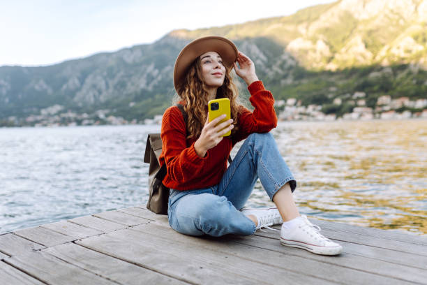 istock A young woman using her phone while sitting by the waterfront in a scenic mountain town during a sunny day. 2174122396
