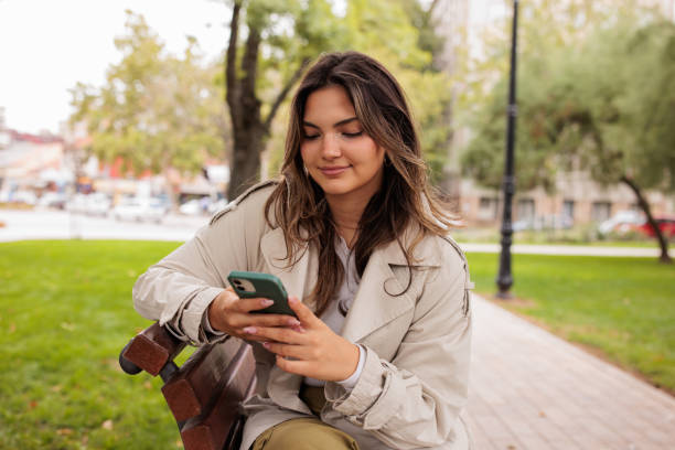 istock Young woman sitting on a park bench and using her phone 2174130156