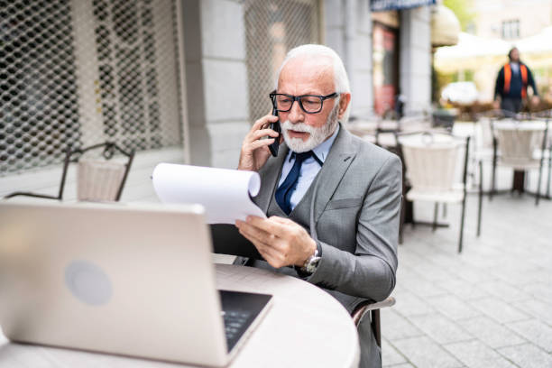istock Modern senior businessman talking on phone and using laptop in cafe 2174136507