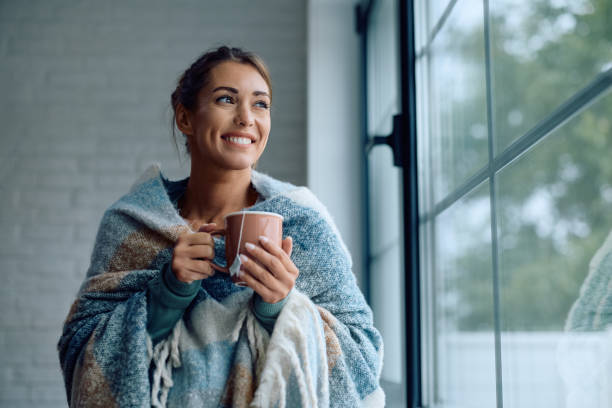 istock Happy woman wrapped in a blanket drinking tea by the window. 2174137143