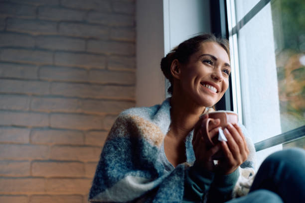 istock Happy woman wrapped in a blanket drinking tea while enjoying by the window at home. 2174137311