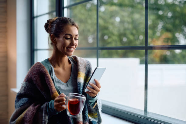 Happy woman wrapped in a blanket drinking tea while using cell phone at home.