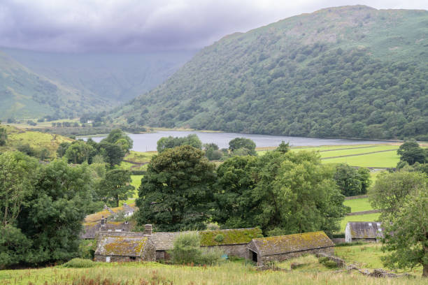 A peaceful countryside scene with rustic stone cottages nestled under trees, set against a backdrop of rolling hills and a tranquil lake. The misty clouds add a dramatic mood to this rural landscape.