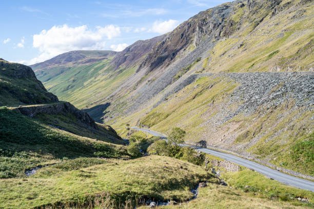 A winding road snakes through rugged mountains, flanked by steep rocky slopes. The afternoon sun highlights the textures of the landscape, offering a breathtaking view of nature's raw beauty and the journey ahead.