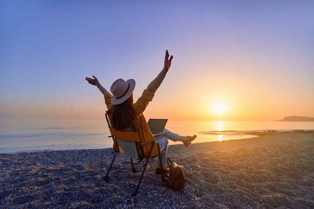 istock Satisfied free millennial freelancer nomad woman with open arms using computer and sitting on the beach by the calm sea. Enjoyment of dream office remote work concept 2174148633