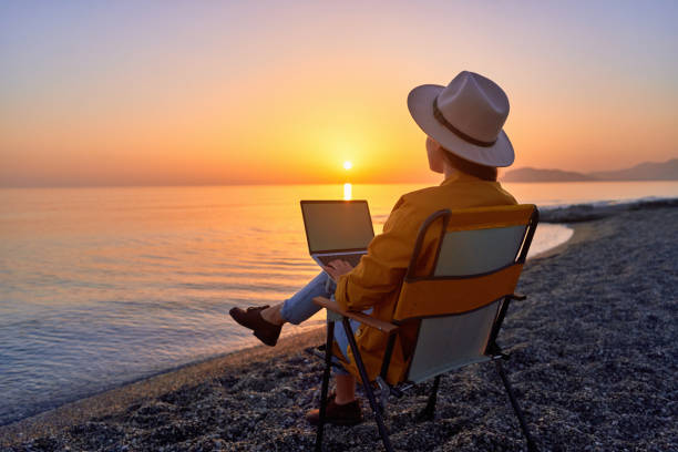istock Free nomad freelancer woman using computer and sitting on beach by calm sea at sunset. Enjoyment of dream office remote work concept 2174148648