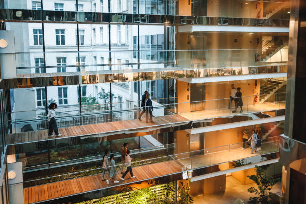 Diverse Professionals Navigate Multi-Level Office Space Men and women in business attire walk through a multi-level office building featuring glass partitions and wooden walkways; modern work setting promoting collaboration and movement. african american architecture stock pictures, royalty-free photos & images