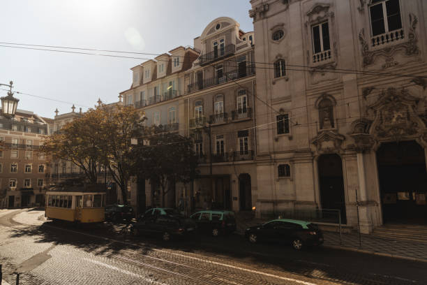 A charming yellow tram crosses a historic street in Lisbon, next to beautiful buildings with classic architecture. The soft sunlight bathes the scene, highlighting the detailed facades and the lines of the tracks. A typical portrait of the bustling and charming urban life in Lisbon.