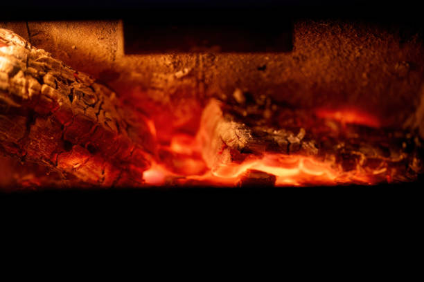 A close-up of firewood burning inside a fireplace, with bright flames and glowing embers. The warm, flickering light highlights the charred wood and creates a cozy atmosphere.