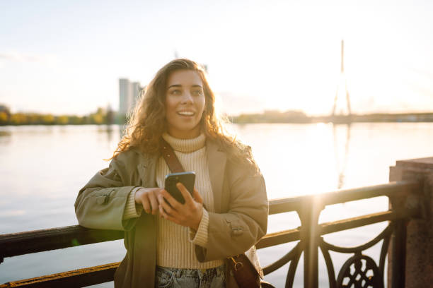 istock Young woman enjoys a sunny day by the river, using her smartphone. 2174156140