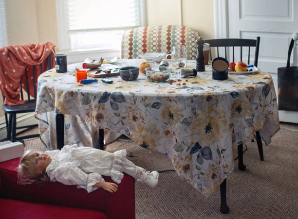 Cluttered dining room table covered with floral tablecloth and various items strewn about, including dishes, food, and household objects. Family appears to be in the midst of moving or unpacking. Doll lies on red surface in foreground. Room has windows with blinds and patterned chair visible in background, adding to domestic disorder.