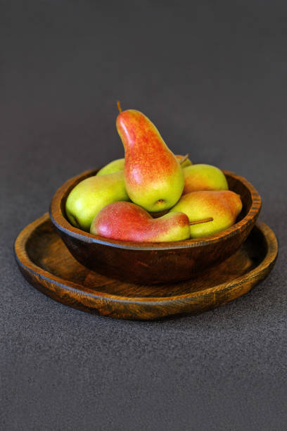 A collection of ripe pears in various shapes of red and green fills a rustic wooden bowl placed on a dark background. This delightful arrangement adds warmth to any kitchen decor.