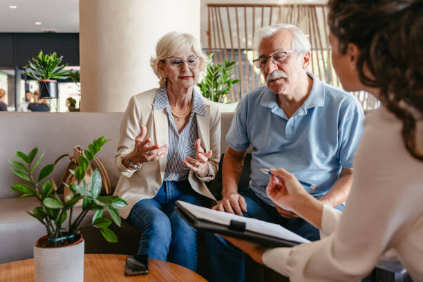 An elderly couple sits in a modern hotel lobby, talking with a hotel staff representative who is assisting them with the check-in process or presenting a hotel offer