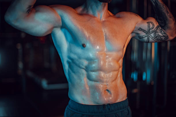 Close-up of a muscular torso with defined abs, tattoos A close-up view of a muscular torso featuring defined abs and tattoos, glistening with sweat in a low-light gym environment, showcasing physical fitness and strength. arm tattoo ideas for men pictures stock pictures, royalty-free photos & images