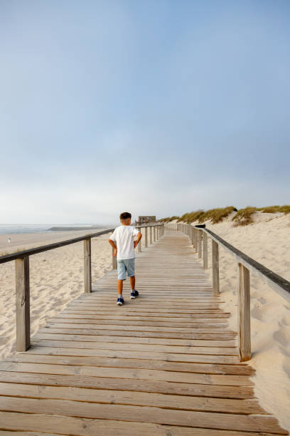 istock A boy walks along a wooden boardwalk on a quiet late afternoon beach with white sand under a cloudy sky, capturing the feeling of freedom and outdoor adventure. 2174172468