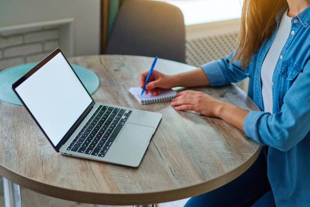 istock Female hands takes a note with pen in a notepad and working at laptop with open white blank display on table at workspace. Computer mockup for online website advertising 2174174607