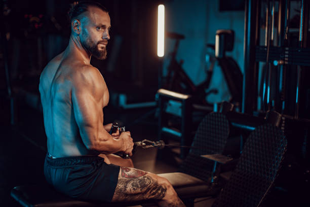 Focused athlete on bench exercising in dark gym A focused, tattooed athlete in a dark gym sits on a bench pulling a cable, embodying strength, determination, and the pursuit of physical excellence. arm tattoo ideas for men pictures stock pictures, royalty-free photos & images