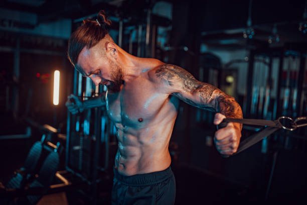 Determined athlete performing cable exercises in gym. A determined, tattooed athlete performs cable exercises, emphasizing his upper body muscles and showcasing the fruits of his dedication in a dimly lit gym. arm tattoo ideas for men pictures stock pictures, royalty-free photos & images