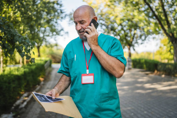 Mature senior man doctor in green medical scrubs stand outdoor, hold envelope with ultrasound picture and talk with patient on mobile phone