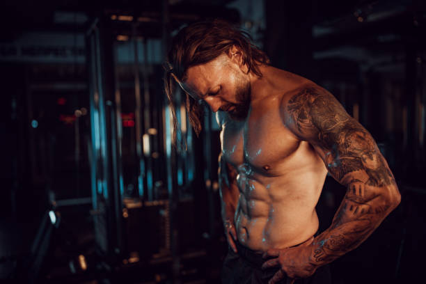 Muscular man with tattoos posing in dark gym area. A muscular man with tattoos on his arms poses shirtless in a dimly lit gym, showcasing his defined muscles and serious expression, highlighting fitness strength. arm tattoo ideas for men pictures stock pictures, royalty-free photos & images