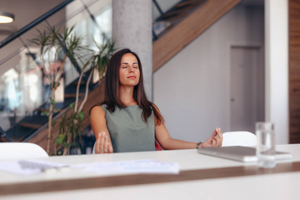Businesswoman Practicing Meditation at Office Desk for Stress Relief A woman in a modern office setting practices meditation at her desk to achieve stress relief and mindfulness. The image conveys calmness and balance in a work environment. meditation room office stock pictures, royalty-free photos & images