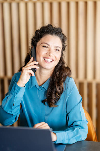istock A cheerful woman wearing a blue blouse is sitting at a table, talking on her phone while smiling. She has a laptop in front of her, appearing to be in a work setting. 2174225427