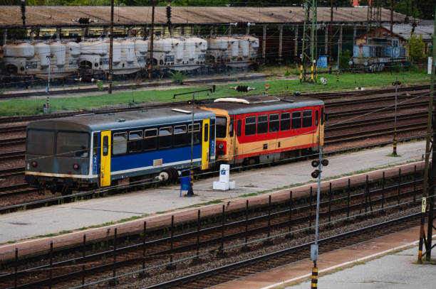 A red and yellow train travels alone on a set of railway tracks amidst a lush green landscape. The scene features multiple tracks, overhead power lines, and steel pylons, reflecting a busy yet organized rail transportation system.