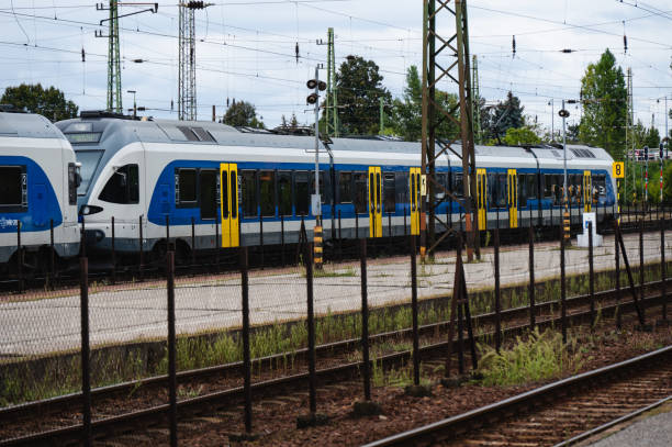 Two modern commuter trains with a blue, white, and yellow color scheme are parked at a railway station. The scene features overhead power lines, green pylons, and fencing, highlighting a contemporary public transportation system in an urban setting.