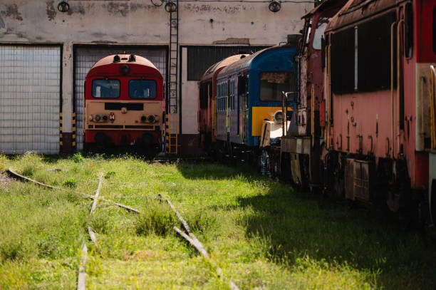 A group of old trains, showing signs of rust and wear, are parked side by side in an abandoned railway depot. The locomotives, in varying shades of red, orange, and blue, contrast with the industrial backdrop and overgrown grass surrounding the area.