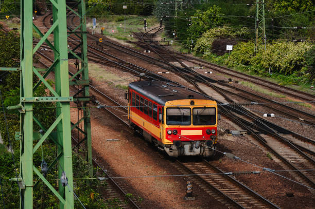 A red and yellow train travels alone on a set of railway tracks amidst a lush green landscape. The scene features multiple tracks, overhead power lines, and steel pylons, reflecting a busy yet organized rail transportation system.