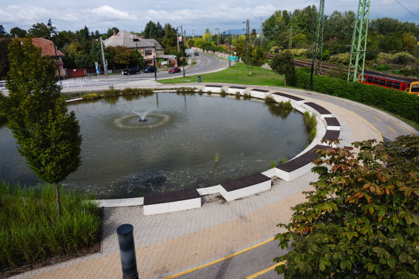 A peaceful circular fountain is surrounded by a paved walkway and benches in a small park. The setting is close to a railway line, with greenery and a quiet suburban street visible in the background, creating a calm urban landscape.