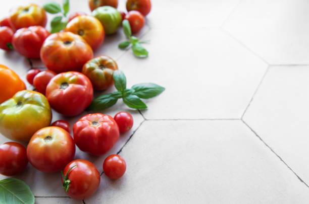 A variety of heirloom tomatoes, including red, yellow, and green, are arranged casually on a light-colored hexagonal tiled surface alongside vibrant basil leaves.