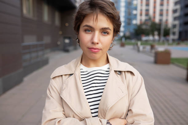 istock Young stylish woman facing housing problems, standing with crossed hands on background with residential area ad multi-storey buildings, looking at camera with serious face, trying to find solution 2174330396