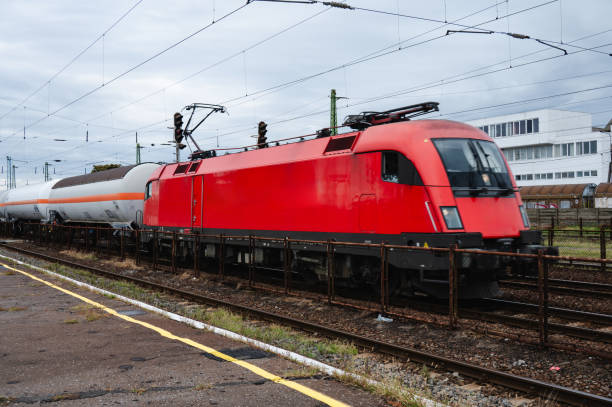 A red freight train pulling a series of tank cars at a railway station, with power lines and signals visible in the background. The industrial scene captures the movement of goods by rail, surrounded by infrastructure and equipment.