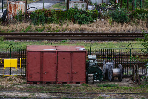 An industrial storage unit with rust-red doors is located near railway tracks, accompanied by cable spools and various machinery. The scene includes overgrown vegetation and rugged infrastructure, reflecting the industrial and functional setting.