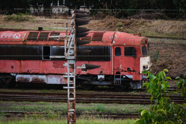 A group of old trains, showing signs of rust and wear, are parked side by side in an abandoned railway depot. The locomotives, in varying shades of red, orange, and blue, contrast with the industrial backdrop and overgrown grass surrounding the area.
