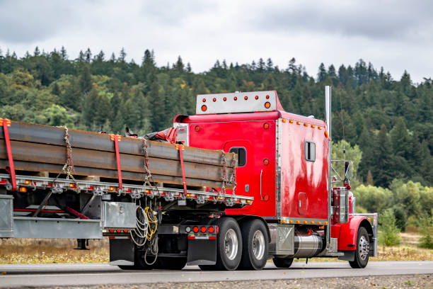 istock Red big rig classic semi truck tractor with extended cab transporting metal profile on flat bed semi trailer running on the highway road with forested hills 2174344412