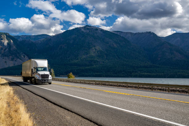Day cab white big rig semi truck with huge roof spoiler transporting cargo in dry van semi trailer running on the highway road along the Columbia River with mountains on the sides stock photo