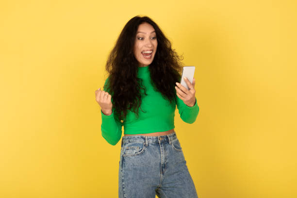 istock Joyful Middle Eastern Lady Holding Cellphone Shaking Fists, Yellow Background 2174345827