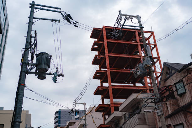 View of a steel-framed structure under construction, rising amidst existing urban buildings. Power lines and transformers are seen in the foreground, showcasing the juxtaposition of industrial development and city infrastructure. The cloudy sky provides a neutral backdrop, emphasizing the sharp lines and reddish hues of the metal frame.
