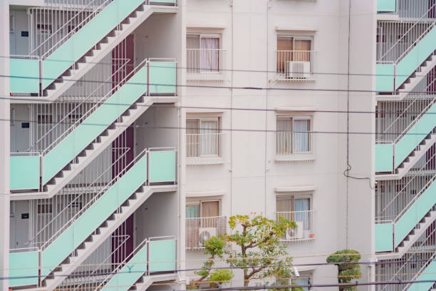 Facade of a modern urban apartment building featuring prominent external staircases and multiple windows. The pastel colors of the staircases contrast with the white walls, creating visual interest. A tree with pruned topiary adds a touch of greenery in the foreground.