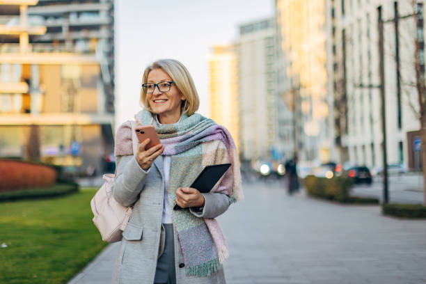 istock A modern and smiling senior woman is using a mobile phone while walking on the sidewalk through the city 2174383755