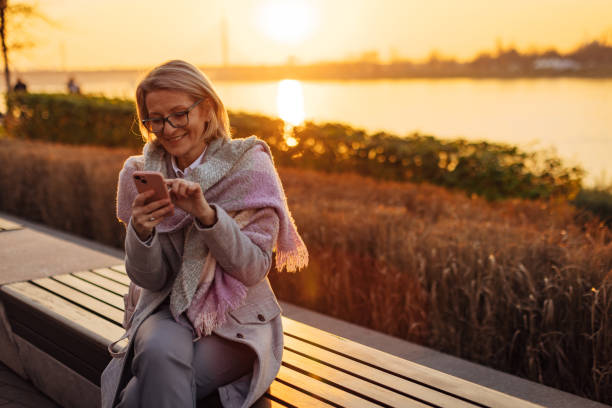 istock Smiling senior adult woman using smartphone while sitting on park bench, overlooking river 2174384383